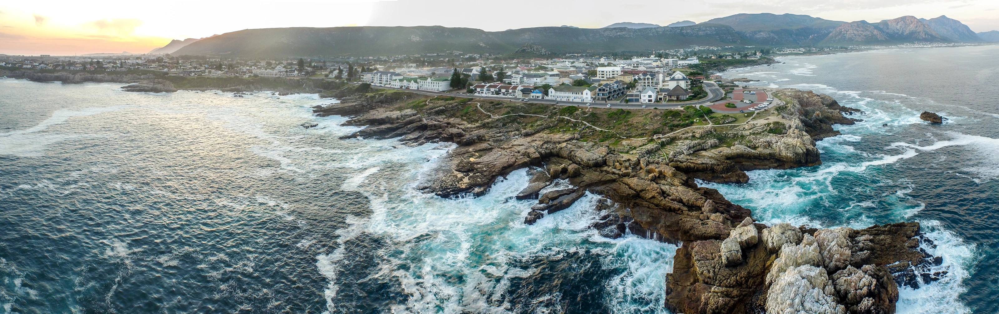 A sweeping aerial view of Hermanus town, rocky coastline and mountains at sunset